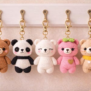 May include: Five handmade crochet bear keychains in brown, panda, white, pink, and tan. Each bear has a gold-colored clasp and is hanging from a white wooden board. The pink bear has a green bow and a heart.
