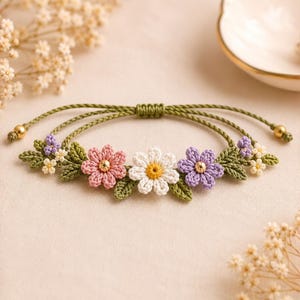 May include: A bracelet featuring a green cord with an adjustable sliding knot closure. The centerpiece displays crocheted flowers in pink, white, and purple, accented with small green leaves and tiny gold-toned beads at the ends of the cord.