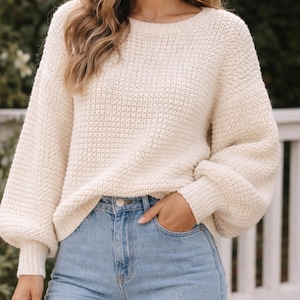 Può includere: Maglione lavorato a maglia color crema con scollo tondo e maniche lunghe e voluminose. Il maglione ha un motivo a maglia strutturato e polsini a coste. La modella indossa jeans azzurri. Il maglione è uno stile casual e confortevole.