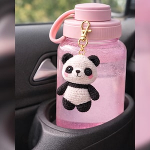 May include: A pink water bottle with a matching lid and handle, held in a car's cup holder. A small, crocheted panda bear charm with black and white details hangs from the bottle. The bottle is filled with clear liquid.