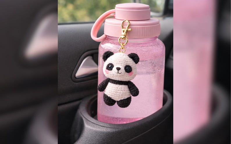 May include: A pink water bottle with a matching lid and handle, held in a car's cup holder. A small, crocheted panda bear charm with black and white details hangs from the bottle. The bottle is filled with clear liquid.