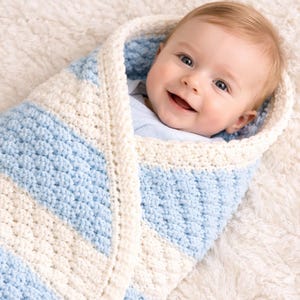 May include: A smiling baby wrapped in a blue and white striped crocheted blanket. The blanket is soft and cozy, with a textured appearance. The baby has blonde hair and is looking directly at the camera.