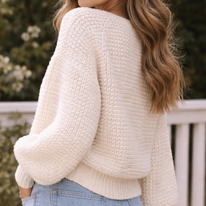 May include: Cream-colored, chunky knit sweater with a wide neckline and long, voluminous sleeves. The sweater has a ribbed hem and cuffs. The person is wearing blue jeans. The background is blurred, with greenery and a white fence.