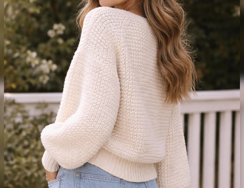 May include: Cream-colored, chunky knit sweater with a wide neckline and long, voluminous sleeves. The sweater has a ribbed hem and cuffs. The person is wearing blue jeans. The background is blurred, with greenery and a white fence.