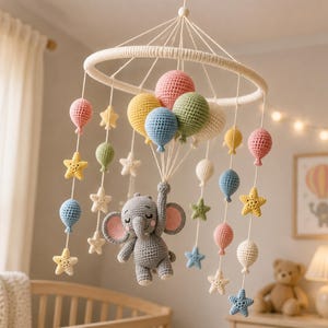 May include: A handmade baby mobile featuring a gray crocheted elephant holding colorful balloons. The mobile includes pink, yellow, blue, and green balloons, along with star-shaped accents. The mobile hangs from a white ring, creating a whimsical nursery decoration.
