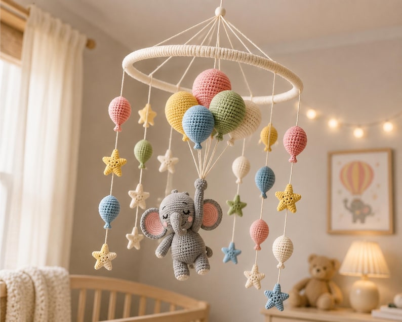 May include: A handmade baby mobile featuring a gray crocheted elephant holding colorful balloons. The mobile includes pink, yellow, blue, and green balloons, along with star-shaped accents. The mobile hangs from a white ring, creating a whimsical nursery decoration.