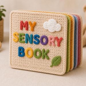 May include: A crocheted sensory book with the words "MY SENSORY BOOK" in colorful letters. The book has a beige cover with a white cloud and green leaf embellishments. The pages are in various colors.