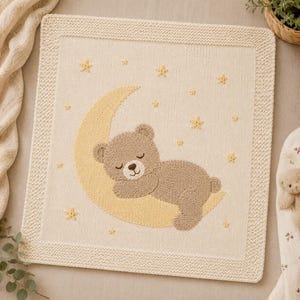 Pode incluir: Uma manta quadrada de malha na cor creme com um design central de um urso de peluche castanho claro a dormir numa lua crescente amarela. O fundo é decorado com pequenas estrelas amarelas bordadas e a borda tem um padrão de malha texturizado.