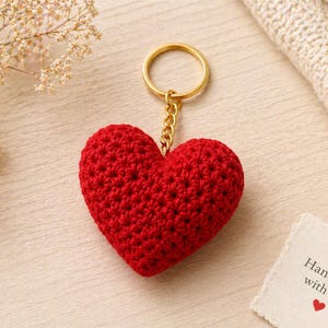 May include: A handmade red crocheted heart keychain with a gold-colored ring and chain. The keychain is on a light wooden surface, with a small card that reads "Handmade with love".