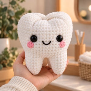 Puede incluir: Un peluche de diente tejido a ganchillo en color blanco con una superficie de hilo texturizado, dos ojos de plástico negro, una boca negra bordada y dos pequeñas mejillas circulares de color rosa. Tiene forma de molar con dos raíces.