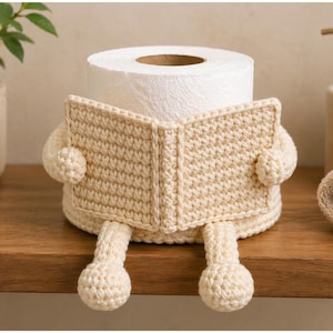 May include: A cream-colored, crocheted toilet paper holder shaped like a person reading a book. The toilet paper roll is the head, with the crocheted book forming the body and arms. The legs are also crocheted.