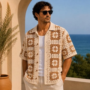 May include: A short-sleeved, button-down crochet shirt in cream and brown. The shirt features a square pattern design and a collared neckline. The person in the image is wearing sunglasses and a white undershirt. The background includes a view of the ocean and a potted plant.