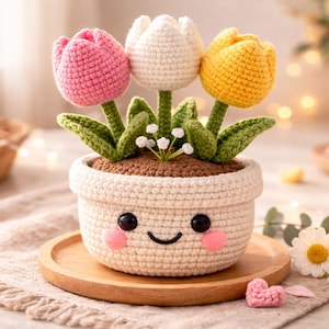Pode incluir: Um vaso de flores de crochê com um rosto sorridente, segurando três tulipas coloridas. As tulipas são rosa, brancas e amarelas. O vaso é creme e está em uma bandeja de madeira. Um pequeno coração rosa está ao lado do vaso.