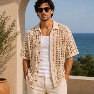 May include: A beige, open-weave crochet shirt with short sleeves and wooden buttons. The shirt is worn over a white tank top and paired with beige shorts. The background features a blue ocean and a beige wall.