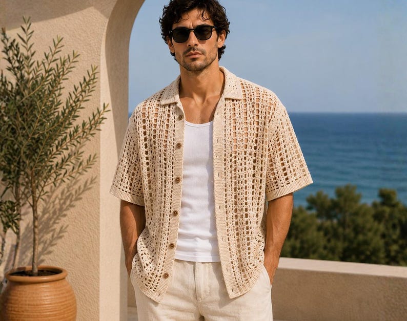May include: A beige, open-weave crochet shirt with short sleeves and wooden buttons. The shirt is worn over a white tank top and paired with beige shorts. The background features a blue ocean and a beige wall.