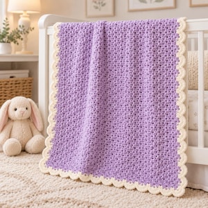 Puede incluir: Una manta de ganchillo rectangular que presenta un centro texturizado de color morado lavanda con un borde festoneado de color crema. El tejido muestra un patrón de puntos repetitivo y consistente en todo el cuerpo, acabado con un borde decorativo.