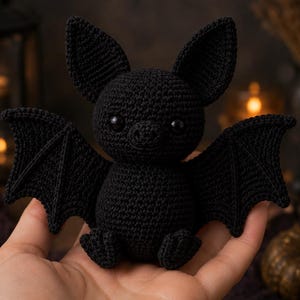 May include: A small, black, crocheted bat plush toy featuring large, pointed ears, wide-set black eyes, and detailed wing structures. The handmade stuffed animal is crafted from dark yarn with a textured, knotted stitch pattern throughout its body.
