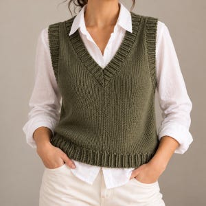 May include: An olive green knit sweater vest featuring a deep V-neckline, ribbed trim along the armholes, neckline, and hem, worn over a crisp white long-sleeved button-down shirt and light-colored pants.