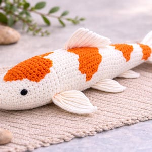 Könnte beinhalten: Ein handgehäkelter Koi-Fisch in Weiß und Orange, der auf einer strukturierten, beigefarbenen Oberfläche liegt. Der Fisch hat detaillierte Flossen und ein schwarzes Auge und zeigt ein einzigartiges, handgefertigtes Design. Dieser Dekorationsartikel ist perfekt für die Inneneinrichtung.