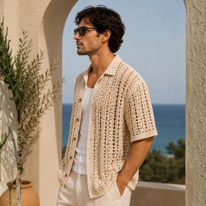 May include: A man wearing a beige crochet shirt with short sleeves and a button-down front, paired with white pants. He is also wearing sunglasses. The background features a view of the ocean and a stone archway, with greenery on the left.