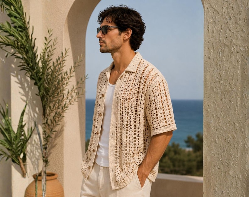 May include: A man wearing a beige crochet shirt with short sleeves and a button-down front, paired with white pants. He is also wearing sunglasses. The background features a view of the ocean and a stone archway, with greenery on the left.