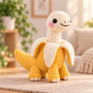 May include: A crochet plush toy shaped like a dinosaur emerging from a yellow banana peel. The creature has a cream-colored head, black button eyes, pink cheeks, and a brown stitched smile. The body is yellow with textured crochet stitches.