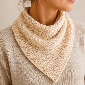 May include: A cream-colored, triangular knit scarf is draped around a person's neck. The scarf is made of a thick, textured yarn and has a simple, classic design. The scarf is a neutral color, making it versatile for various outfits.
