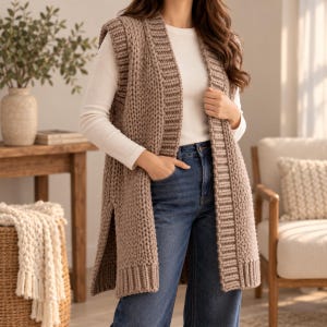 May include: A long, sleeveless, chunky knit vest in a neutral taupe color. The vest features a thick ribbed trim along the front opening, armholes, and hem, with deep side slits and a prominent vertical knit texture throughout the garment.