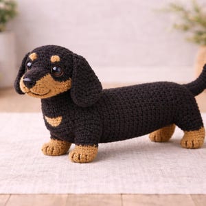 Puede incluir: Un perro salchicha de ganchillo en negro y marrón claro. El juguete de peluche hecho a mano tiene un cuerpo largo, patas cortas y orejas caídas. El perro mide aproximadamente 30 cm de largo y está sobre una superficie neutra.