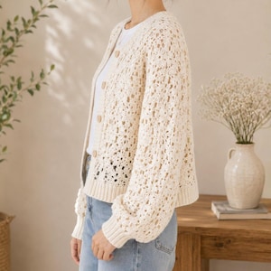 May include: Cream-colored, open-weave cardigan with a button-up front. The long-sleeved sweater has a round neckline and a slightly oversized fit. The cardigan is paired with a white top and blue jeans. The background includes a wooden table and vases with flowers.