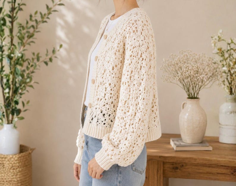 May include: Cream-colored, open-weave cardigan with a button-up front. The long-sleeved sweater has a round neckline and a slightly oversized fit. The cardigan is paired with a white top and blue jeans. The background includes a wooden table and vases with flowers.