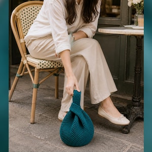 May include: A teal blue, knitted handbag with a knotted handle. The round bag is sitting on a stone surface. The bag is held by a person sitting in a wicker chair.