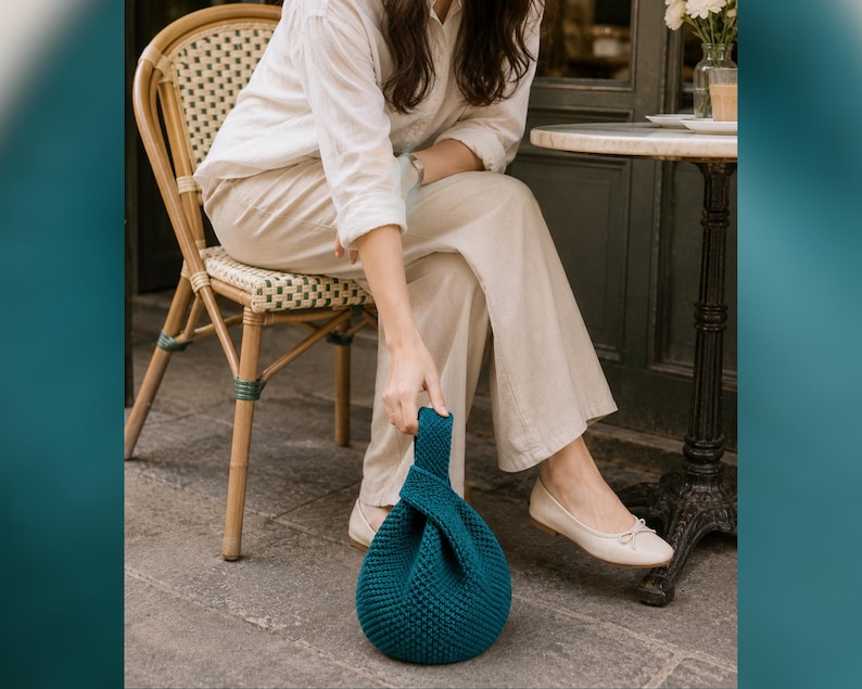 May include: A teal blue, knitted handbag with a knotted handle. The round bag is sitting on a stone surface. The bag is held by a person sitting in a wicker chair.