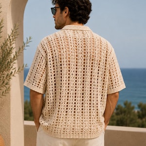 May include: A short-sleeved, open-weave shirt in a light beige color. The shirt has a collared neckline and a loose, breathable design. The image shows the back of the shirt, with the ocean visible in the background.