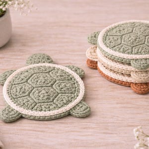 May include: Crocheted turtle-shaped coasters in green, cream, and brown. The coasters feature a textured, hexagonal pattern and a white border. A stack of coasters is visible, along with a single coaster and a vase of white flowers.