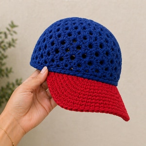 May include: A handmade crochet baseball cap with a blue crown and a red brim. The blue crown has a perforated design. The cap is held by a person's hand.