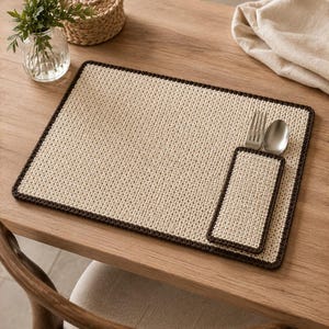 Puede incluir: Un mantel individual rectangular de ganchillo color crema con un borde marrón oscuro. El mantel cuenta con un bolsillo rectangular unido en el lado derecho, que contiene un tenedor de metal plateado y una cuchara de metal plateado.