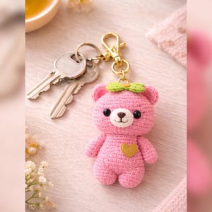 May include: A pink crocheted bear keychain with a gold-colored heart and a green bow. The bear has black eyes and a brown nose. Two keys are attached to the keychain. The background is a soft pink.