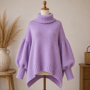 May include: A chunky knit jumper in a light lavender colour featuring a high turtleneck collar, long voluminous balloon sleeves with ribbed cuffs, and a high-low hemline. The garment is displayed on a mannequin, showcasing the thick yarn texture.