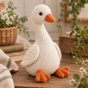 Op de afbeelding: Een handgebreid knuffelgans-speeltje met een crèmekleurig lichaam van gestructureerd garen, een feloranje snavel en bijpassende oranje zwemvliezen. De gans heeft kleine zwarte kraaloogjes en is vormgegeven met een zacht, rond silhouet.