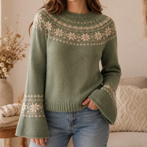 May include: A sage green long-sleeve sweater featuring a cream-colored snowflake pattern around the round neckline and on the flared sleeve cuffs. The knit garment has a ribbed crew neck, ribbed hem, and wide bell-shaped sleeves with a detailed design.