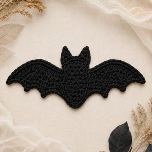 May include: A black crochet bat applique featuring a central body with two pointed ears and wide, scalloped wings. The bat is crafted with a textured yarn stitch pattern, displaying a flat, symmetrical silhouette with detailed wing edges.