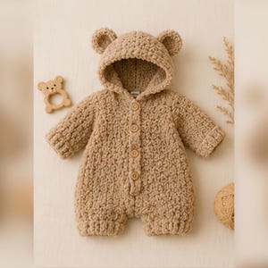 May include: A tan-colored, crocheted baby romper with a bear hood and ears. The romper has long sleeves, button closures, and short legs. A wooden bear-shaped teether is next to the romper.