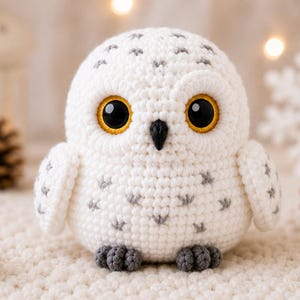 May include: A round, crocheted snowy owl figurine featuring white yarn with small grey accents, large black eyes with yellow irises, and a small black beak. The owl has two small grey feet at the base and a soft, textured, handmade appearance.