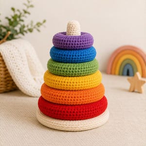May include: A crochet stacking toy featuring a series of six colorful rings in purple, blue, green, yellow, orange, and red, arranged on a cream-colored base with a matching cream-colored post and a small rounded topper at the very peak.