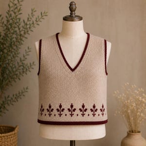Puede incluir: Un chaleco de punto beige con cuello en V y un motivo floral decorativo. El chaleco tiene un ribete burdeos alrededor del escote, las sisas y el dobladillo. El motivo floral también es burdeos.