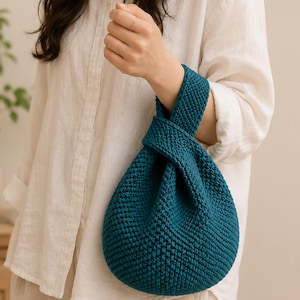 May include: Teal-colored knitted bag with a knotted handle. The bag has a rounded shape and a textured surface. The handle is wide and appears to be made of the same material as the bag. The bag is held by a person wearing a white shirt.