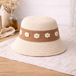 May include: A cream-colored crochet bucket hat with a brown band adorned with white daisy appliqués. The hat has a wide brim and a rounded crown. This sun hat is perfect for spring and summer.