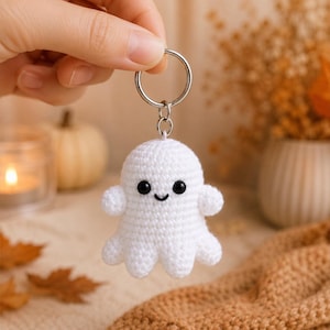 May include: A white crocheted ghost keychain featuring a rounded head, two small arms, a scalloped bottom edge, and two black bead eyes with a stitched black mouth, attached to a silver-colored metal split ring by a small jump ring.