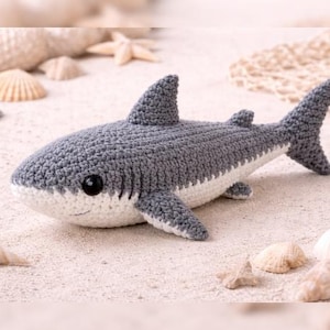 May include: A crocheted shark toy in shades of gray and white, with black safety eyes and a smiling mouth. The handmade shark is lying on a sandy surface, surrounded by seashells and a starfish, evoking a beach theme.